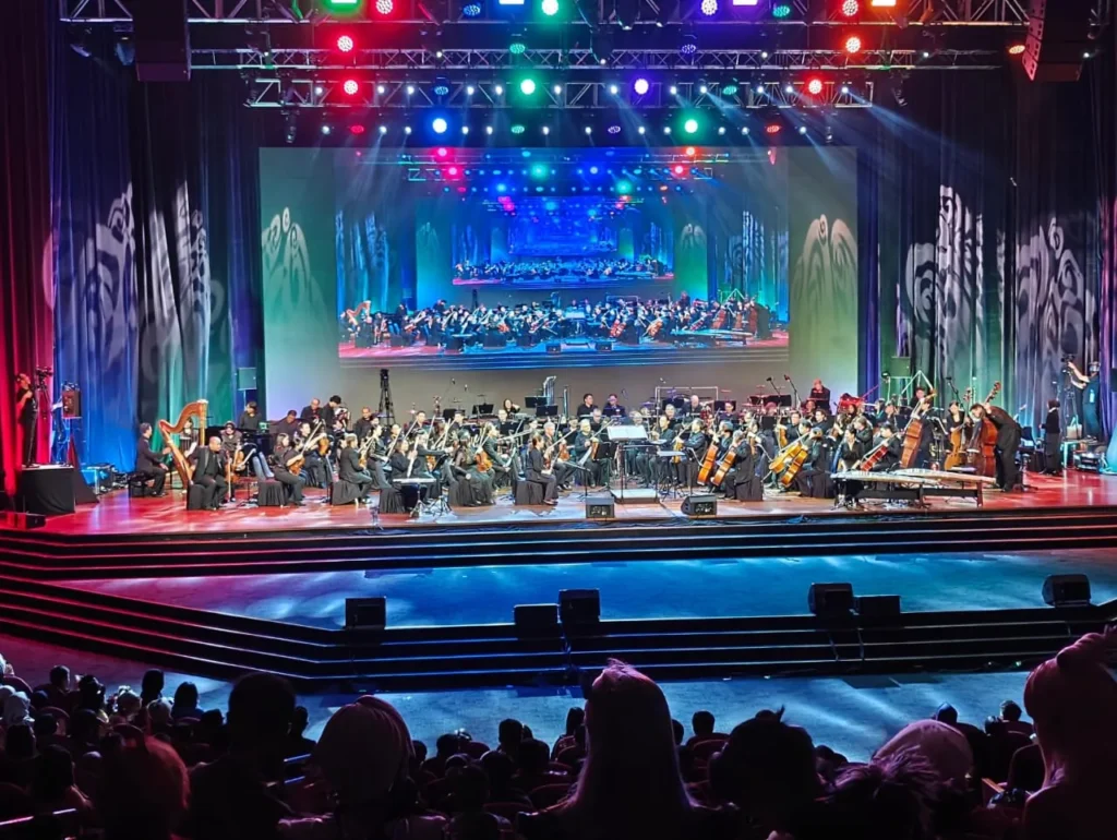 Orchestra on the Auditorium stage with coloured lights and a screen above