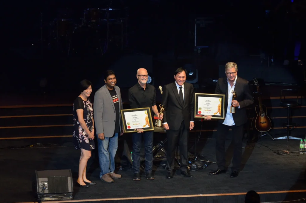 Two award winners holding framed certificates and trophies beside three others on the Auditorium stage