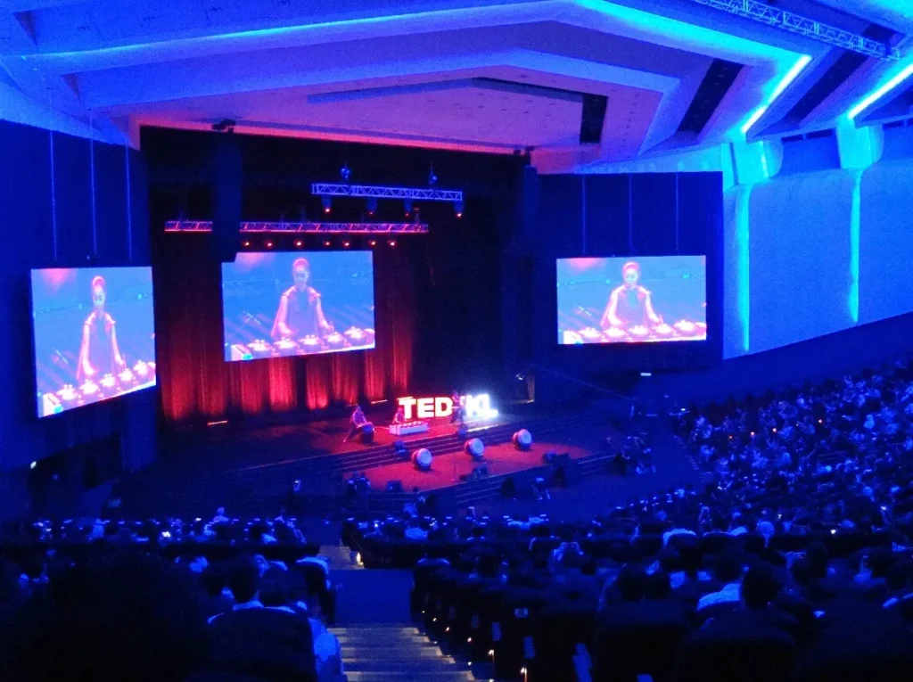Audience watching a TEDxKL talk in the Auditorium, with the speaker on three screens