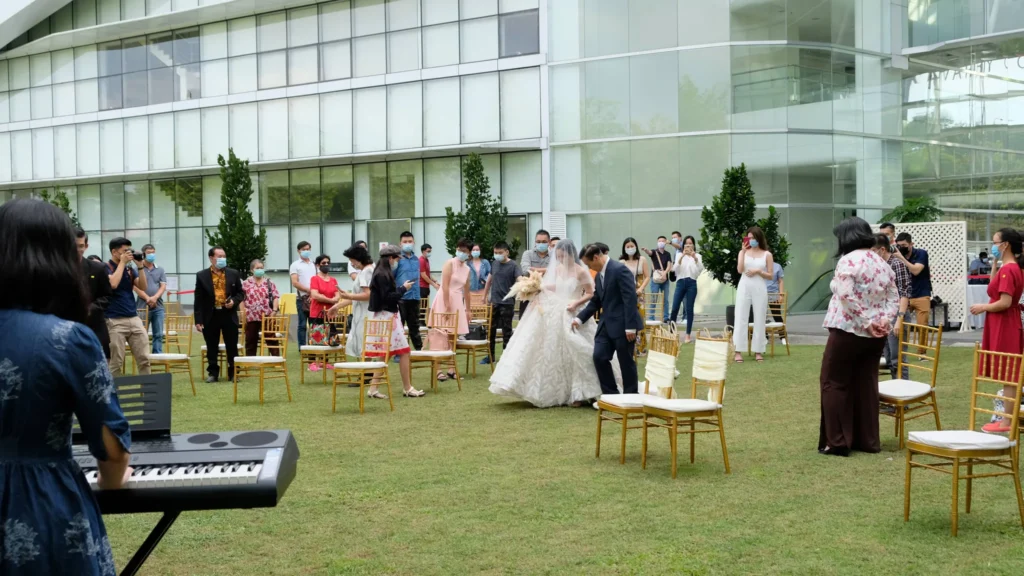 Veiled bride walking in on a man's arm between standing guests on the Garden lawn