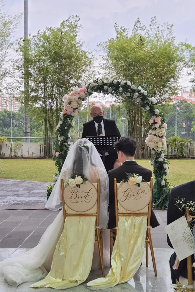 Bride and groom seated on chairs marked Bride and Groom in front of a floral arch in the Garden