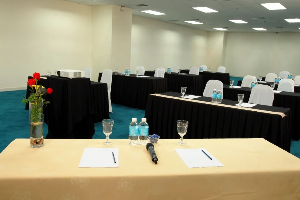 Head table with a microphone, notepads and flowers facing classroom-style tables in a meeting room
