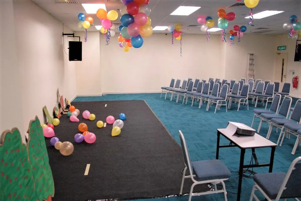 Performance area with balloons and a projector table beside rows of chairs in a meeting room