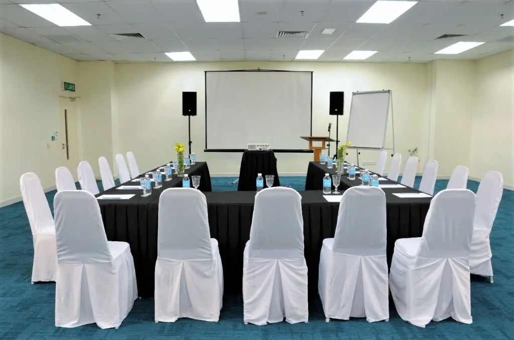 Tables in a U-shape with white chair covers facing a projector screen in a meeting room