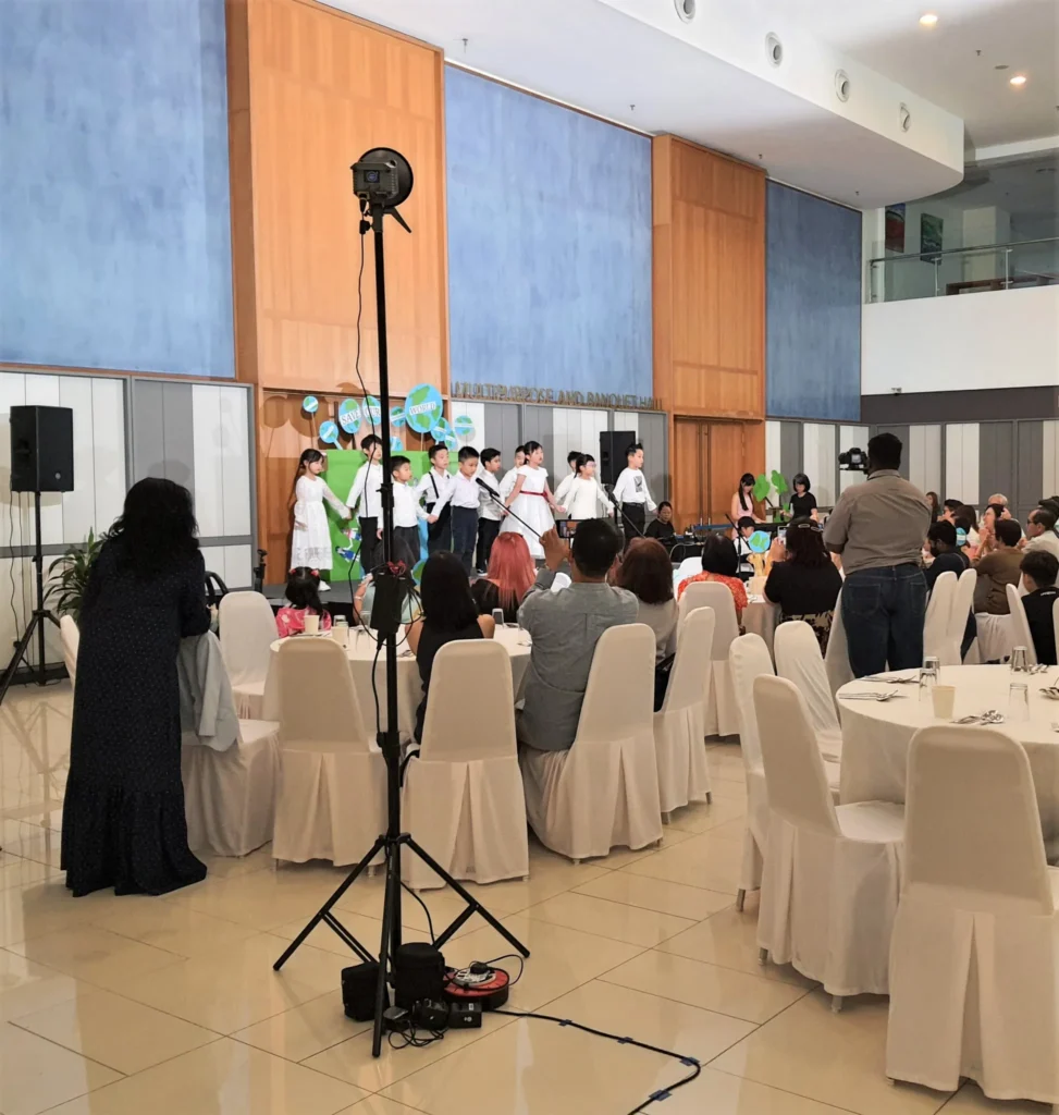 Families at round tables watching a kindergarten graduation on stage in the Multipurpose Banquet Hall lobby