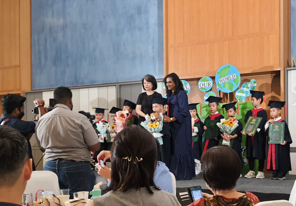 Kindergarten graduates in gowns holding flowers on stage in the Multipurpose Banquet Hall lobby