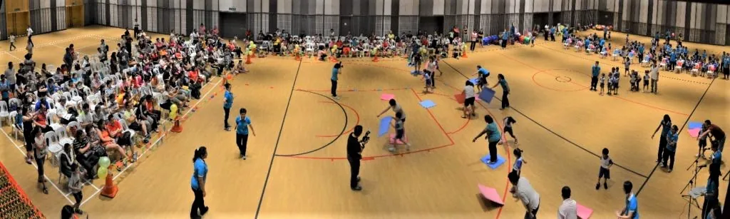 Children playing games at a kindergarten sports day in the Multipurpose Banquet Hall, watched by families