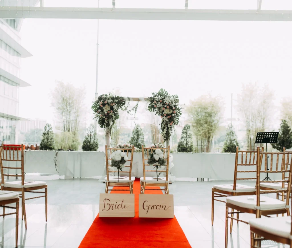 Red carpet aisle leading to a floral arch, with Bride and Groom signs on two chairs in the Plaza