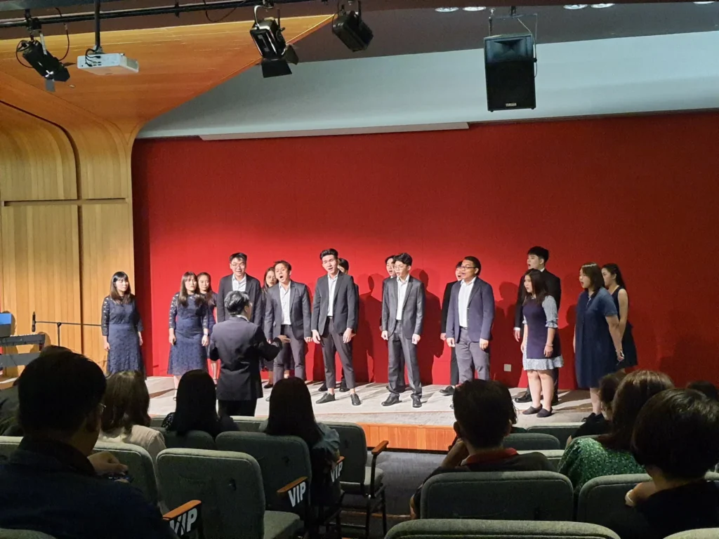 Choir in dark suits and dresses singing at the Elysian Concert in a Theatrette
