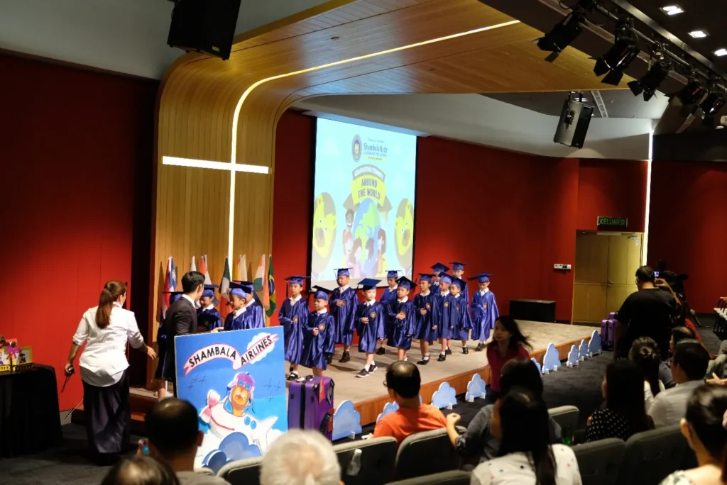 Kindergarten graduates in blue gowns lined up on a Theatrette stage