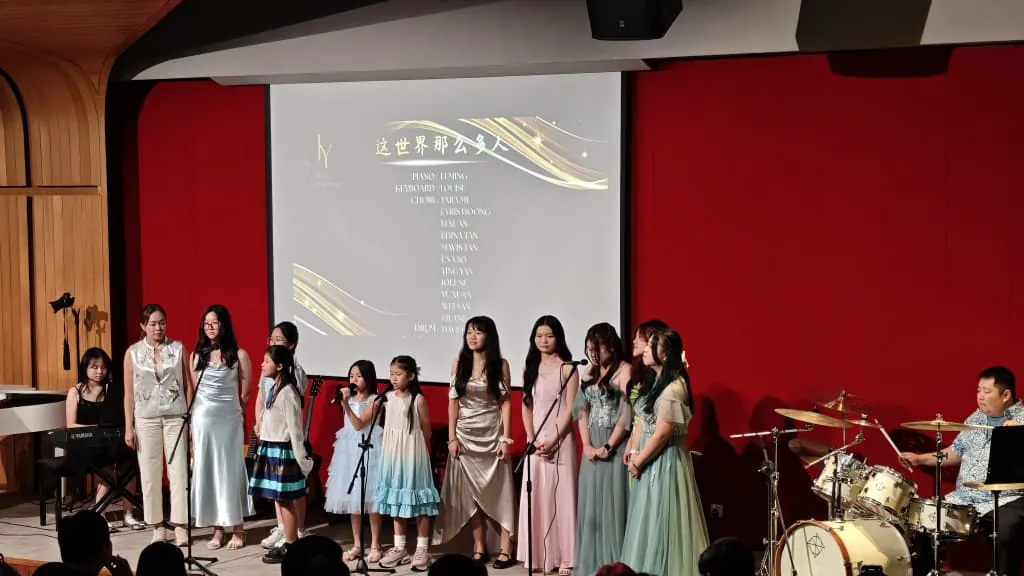 Girls in dresses singing with a pianist and drummer at a music academy recital in a Theatrette