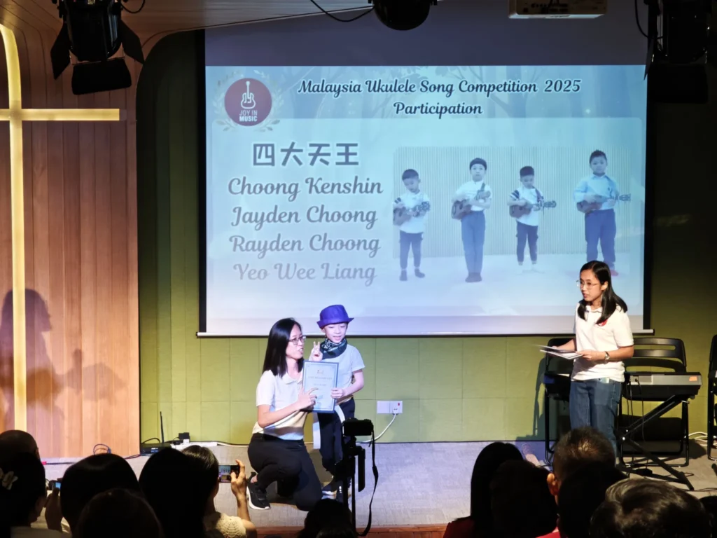 Child receiving a certificate on a Theatrette stage below a ukulele competition slide